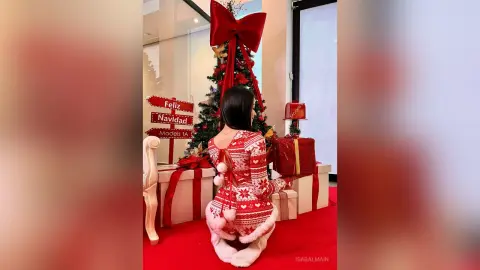 Woman in red festive sweater sitting by a decorated Christmas tree and gifts in a cozy holiday setting.