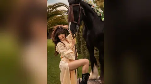 SophiaNapoles poses outdoors next to a black horse, wearing a light yellow dress and showing off her long brown hair and legs.