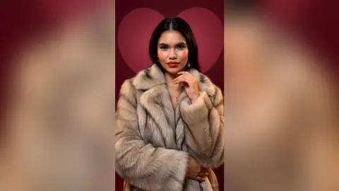 Elegant woman in a luxurious beige faux fur coat posing against a romantic red heart background.