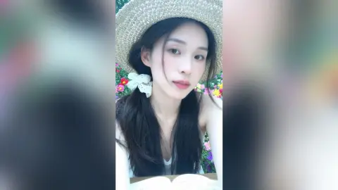 ZhiYue wears a straw hat and a white top, posing with her long dark hair down against a background of colorful flowers.