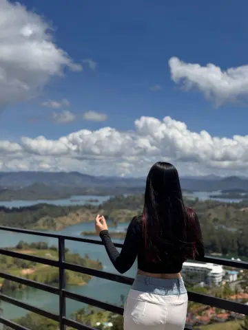 Woman in black long-sleeve shirt and white pants looking at scenic mountain lake view from a balcony.