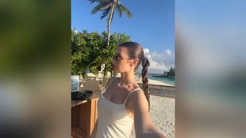 Young woman in a white tank top and sunglasses relaxing on a sunny tropical beach near palm trees.