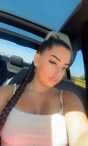 Young woman with long dark braid in white tank top sitting in a car under a sunny blue sky.