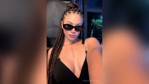 Stylish woman with long braids and sunglasses in a black tank top posing in a moody nighttime setting.