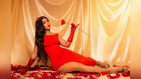 Elegant woman in a red cocktail dress holding a single rose against a gold draped backdrop.