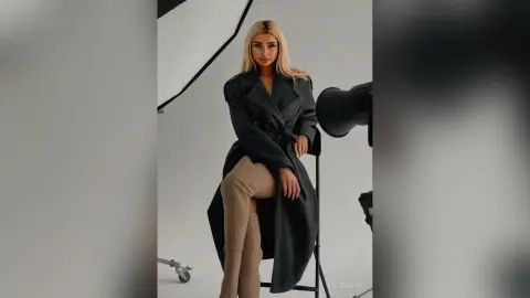 Blonde woman in a stylish black trench coat and beige trousers posing in a professional photo studio.
