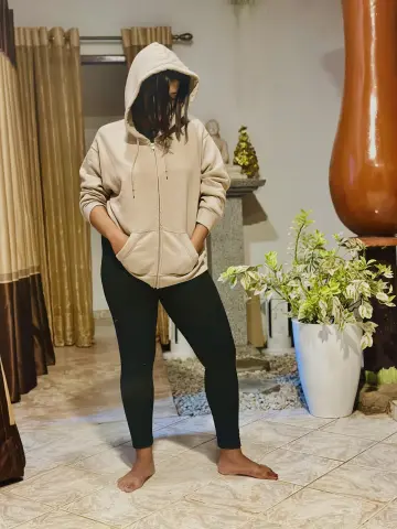 Angel_Myurs — Angel_Myurs stands in a room wearing a beige zip-up hoodie and black leggings with her hands in her pockets.