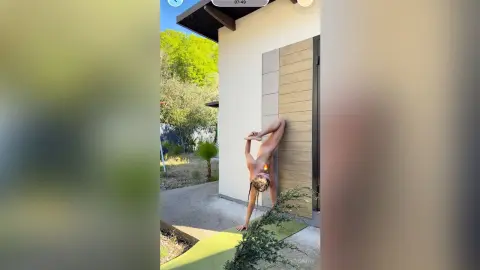 Fit woman in a leopard print bikini performing a standing yoga stretch on a patio near lush green trees.