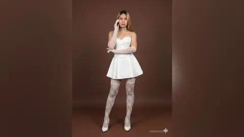 Elegant woman in a white mini dress and lace gloves posing against a brown studio background.