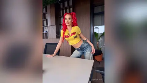 Red-haired woman in a yellow crop top and denim jeans leaning on a table in a modern outdoor patio setting.