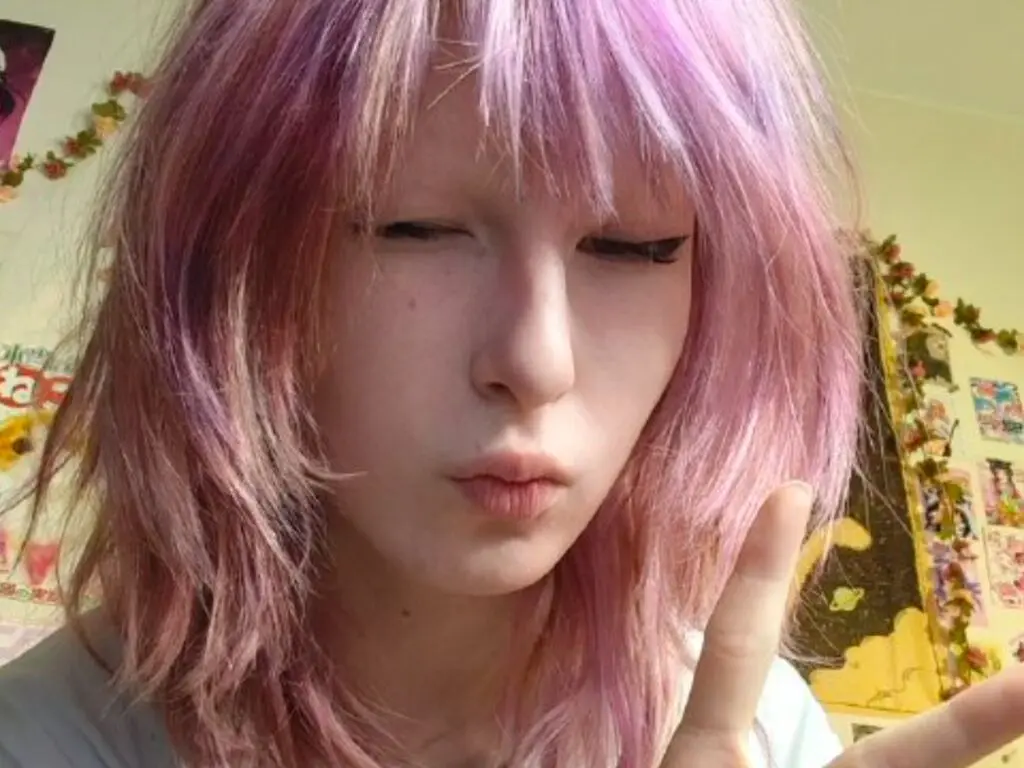 Young woman with pastel pink hair and winged eyeliner making a peace sign in a bedroom setting.