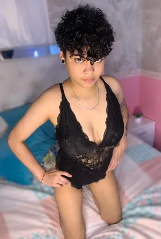Young woman with curly hair wearing a black lace bodysuit sitting on a bed in a bright bedroom.