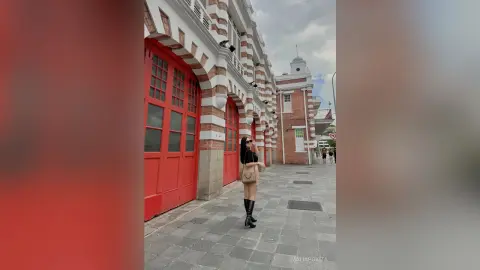 Stylish woman in black outfit and boots standing by red doors of a historic brick building on a city street.