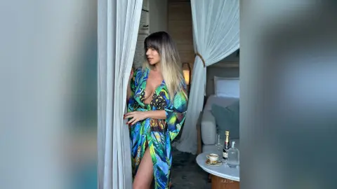 Katia poses in a colorful patterned robe, peeking through white curtains in a bedroom setting.