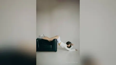 Fashionable woman in white outfit stretching on a black leather sofa in a minimalist studio setting.
