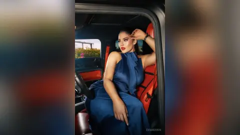 Elegant woman in a navy blue satin dress sitting inside a luxury car with red leather interior.