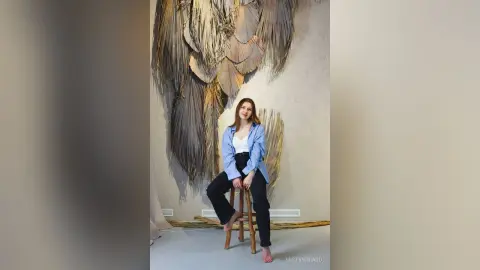 Smiling young woman in a blue button-down shirt and black trousers sitting on a wooden stool in a modern studio.