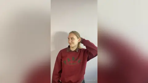 Smiling blonde woman in a burgundy oversized graphic sweatshirt posing against a plain neutral wall.