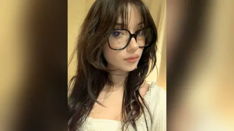 SabrinaSteels poses with long dark hair and large black glasses, wearing a white top against a plain background.