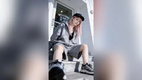 Young woman in grey hoodie and black cap sitting on urban concrete stairs in a casual streetwear style.