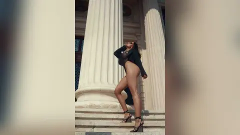 NicoleBella poses against a large stone column wearing a black blazer and black strappy heels.