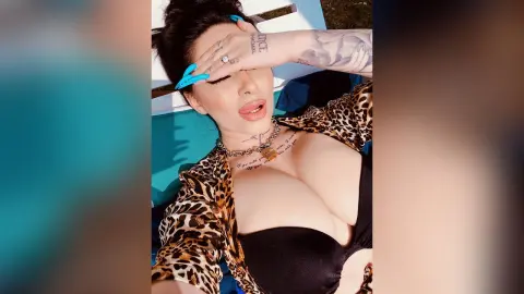 DeeaAnderson reclines in a leopard print top, showing cleavage and her tattoos while resting her hand against her forehead.