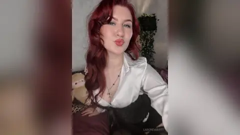 Redheaded woman in a white satin blouse and black lace top smiling in a cozy indoor setting.