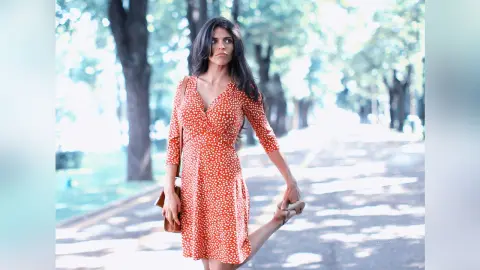 Elegant woman in a red polka dot dress posing in a sunny park setting with a dreamy mood.