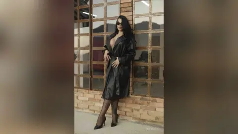 Stylish woman in a black leather trench coat and sunglasses posing confidently by a large glass window.