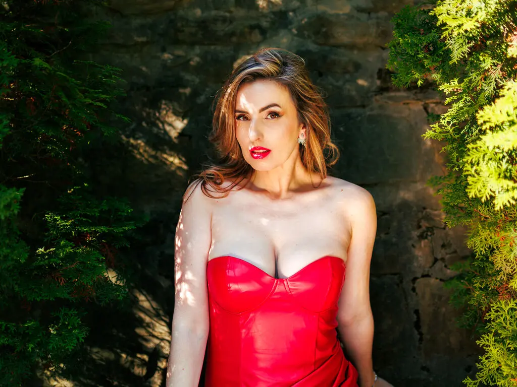 Elegant woman in a red strapless dress posing against a stone wall in a sunny garden setting.