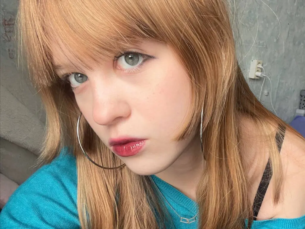 Blonde woman with bangs and silver hoop earrings wearing a teal top in a casual indoor setting.