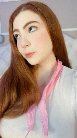 Redheaded woman in a white blouse with a pink ribbon collar posing in a bright, soft-lit indoor setting.