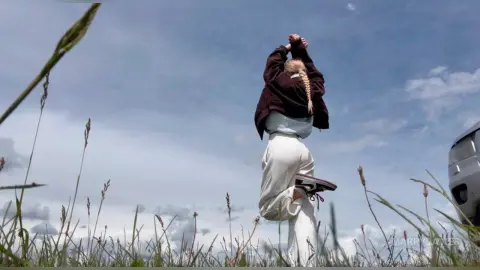 MarryDavies stands in a grassy field with her arms raised above her head, wearing a dark brown long-sleeve top and white pants.