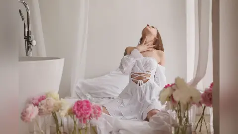 IngridBeckton poses in a white lace outfit, arching her back and touching her neck while sitting on a bed surrounded by pink and white flowers.