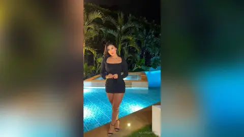 Elegant woman in a black mini dress posing by a luxury swimming pool at night under tropical palm trees.