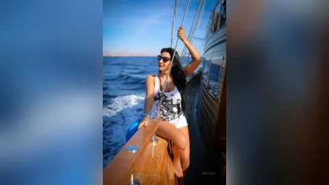 MissyJolie sits on the wooden deck of a boat wearing a white tank top and white shorts, looking out over the blue ocean.