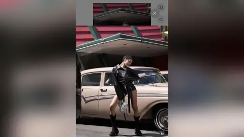 Stylish woman in a black leather jacket and shorts posing next to a vintage car in an urban setting.