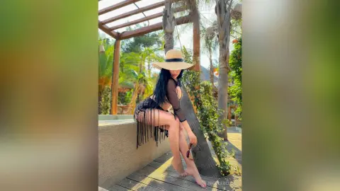 NicoleBella sits on a ledge wearing a black fringe bodysuit and a wide-brimmed straw hat outdoors.