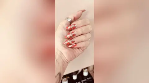 Close-up of a hand with long red almond-shaped nail art and silver rings against a neutral background.