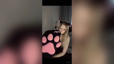 Blonde woman in a cozy black outfit holding a large pink paw plushie in a dimly lit room.
