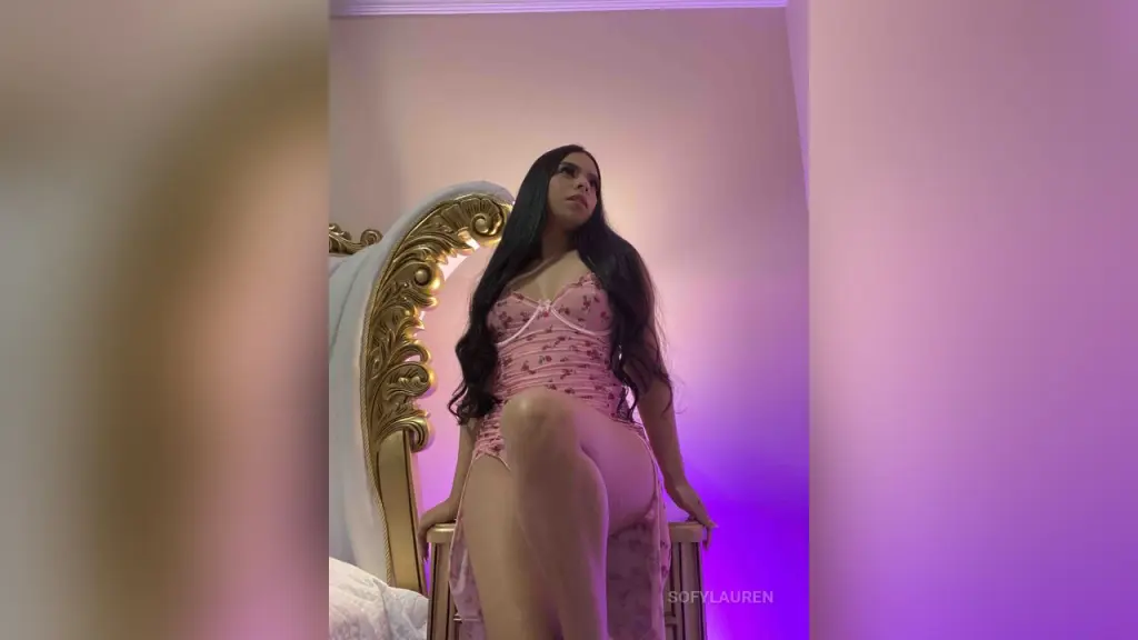 SofyLauren — Young woman in a pink floral lace bodysuit sitting on a gold ornate bed in a moody purple lit bedroom.
