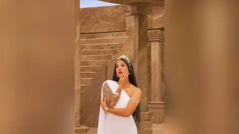 JuliahLewis poses in a white off-the-shoulder top and gold headpiece against a stone staircase background.