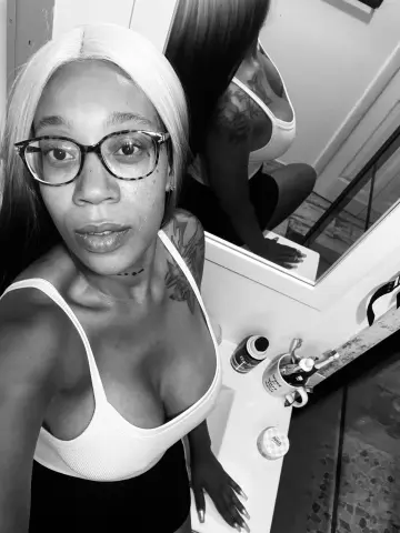 LexBright wearing glasses and a white tank top, posing in front of a bathroom mirror.