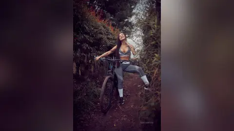 Smiling woman in grey athletic wear posing with a bicycle in a lush, moody forest setting.