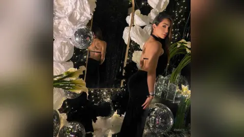 KimRece poses in a black halter top and black pants, standing next to a disco ball and white flowers.