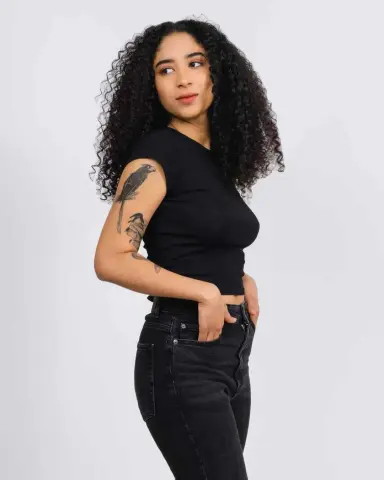 Young woman with curly hair wearing a black t-shirt and dark jeans posing against a white background.
