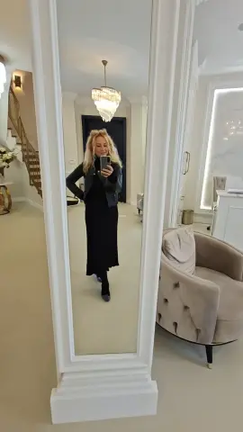 Blonde woman in a black dress and denim jacket taking a mirror selfie in a bright, modern living room.