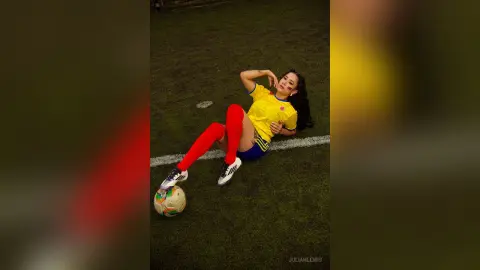 JuliahLewis reclines on a green field wearing a yellow jersey, blue shorts, and long red socks with a soccer ball at her feet.