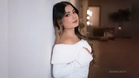 JessieOlivetti poses against a white wall wearing a white off-the-shoulder top with a white faux-fur trim.