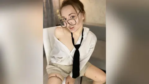 Young woman in glasses and white shirt with loose black tie sitting in a bright room with a playful mood.